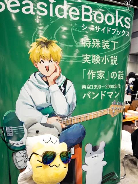 写真:2025/11/23 文学フリマ東京41 カシマのポスターの前でにゃんぷっぷー(blobcat)ぬいぐるみを掲げるねずみちゃん